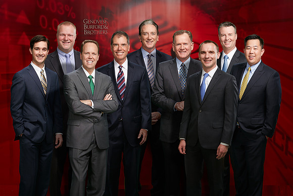 9 executives in suits stand smiling as group in front of a red Genovese Burford and Brothers's branded background